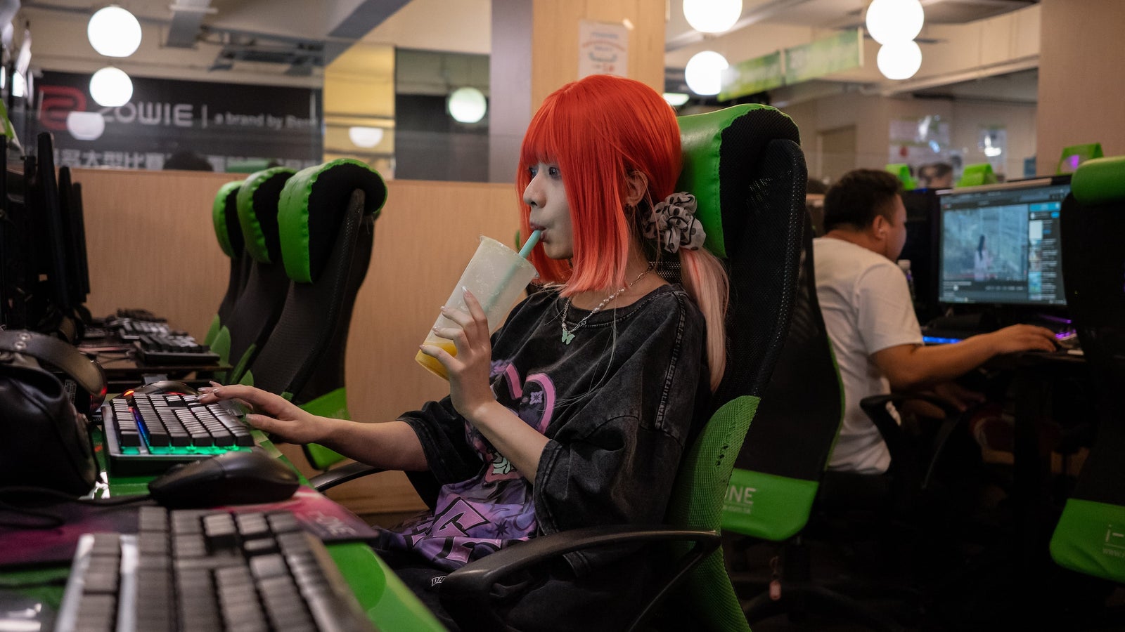 A photo of a woman with red hair drinking from a cup with a straw while seated at a computer in an internet cafe.