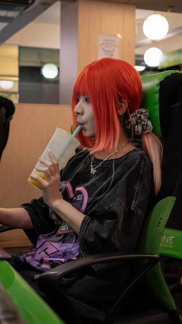 A photo of a woman with red hair drinking from a cup with a straw while seated at a computer in an internet cafe.