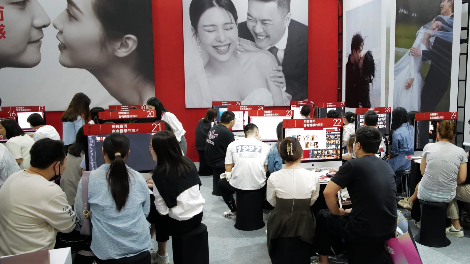 A photo taken from behind of couples seated at computer kiosks, against a backdrop of large-sized wedding photos.