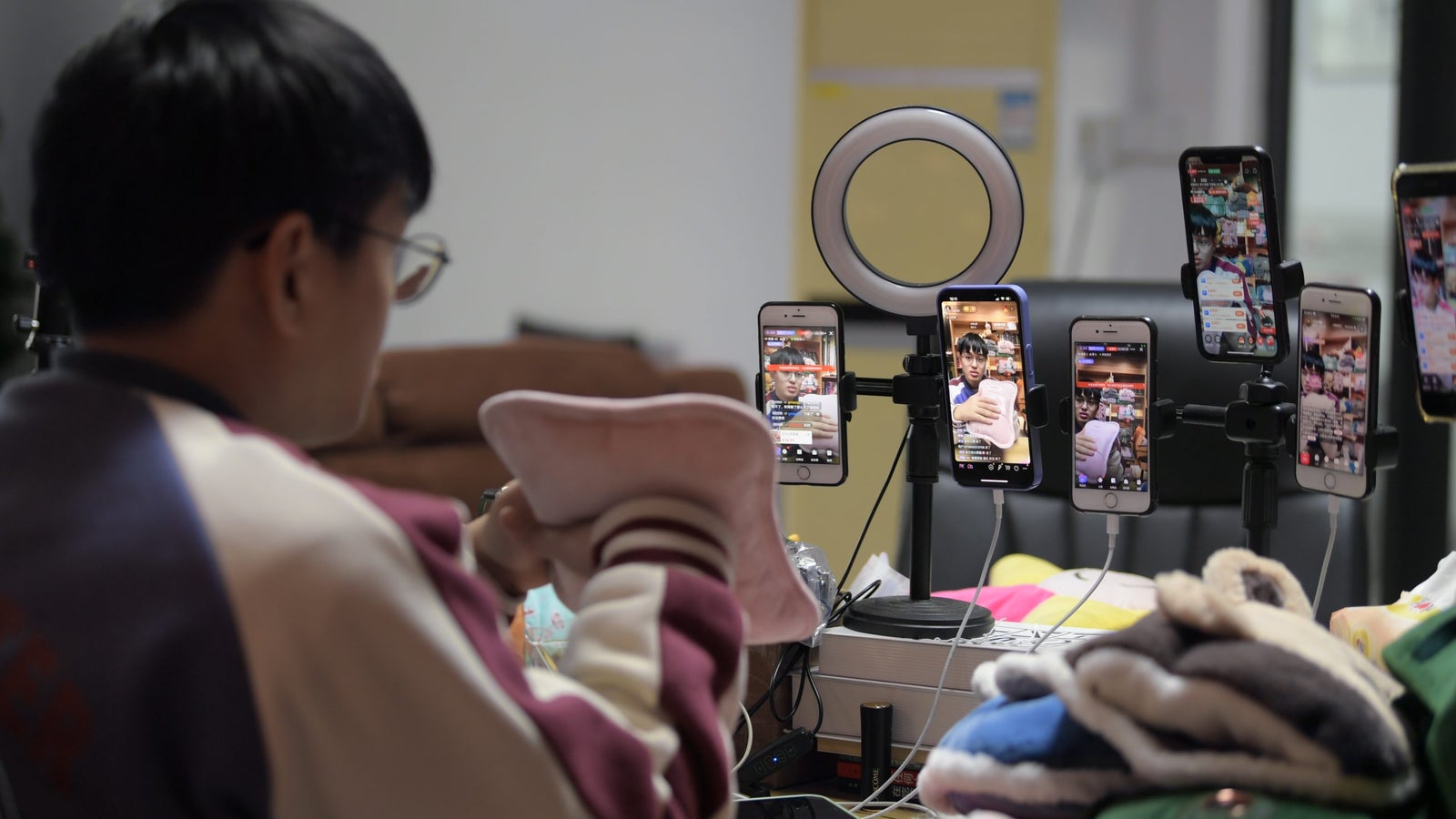 A man holds an electric hot water bottle in front of a livestream recording set up and multiple phone screens.