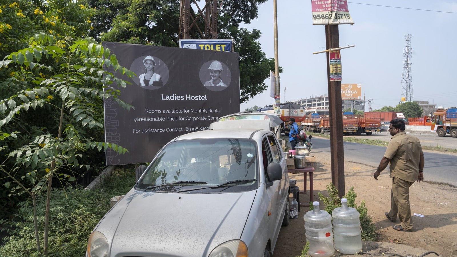 A photo showing a street scene of a parked car, a billboard advertising a "Ladies Hostel", water jugs, a street, and a man walking by.
