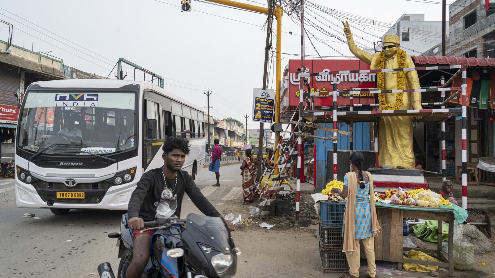 A photo showing a street scene in India with a man riding a motorbike, a golden idol draped in flowers with his hand raised, a bus, and buildings with telephone lines running between them.