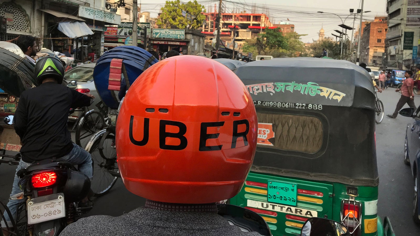 A motorbike rider wearing an orange Uber helmet photographed from the back, on a crowded street in Dhaka, Bangladesh.