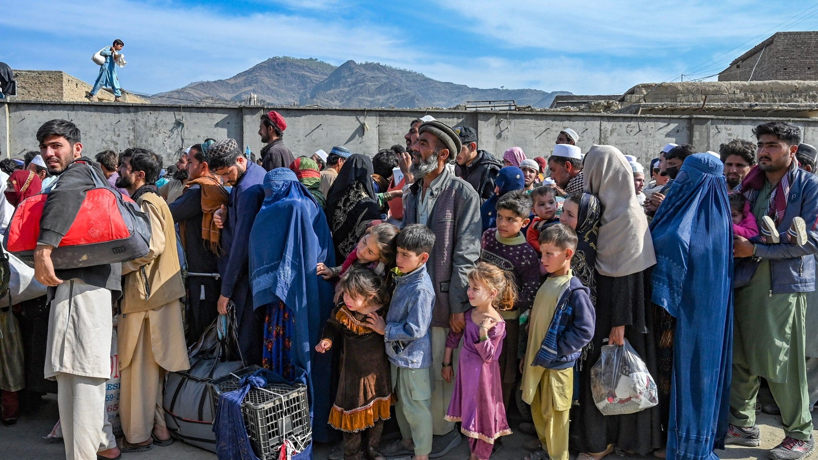 A photo showing a group of Afghan refugees waiting in a line with a blue sky, wall, and mountains in the background.
