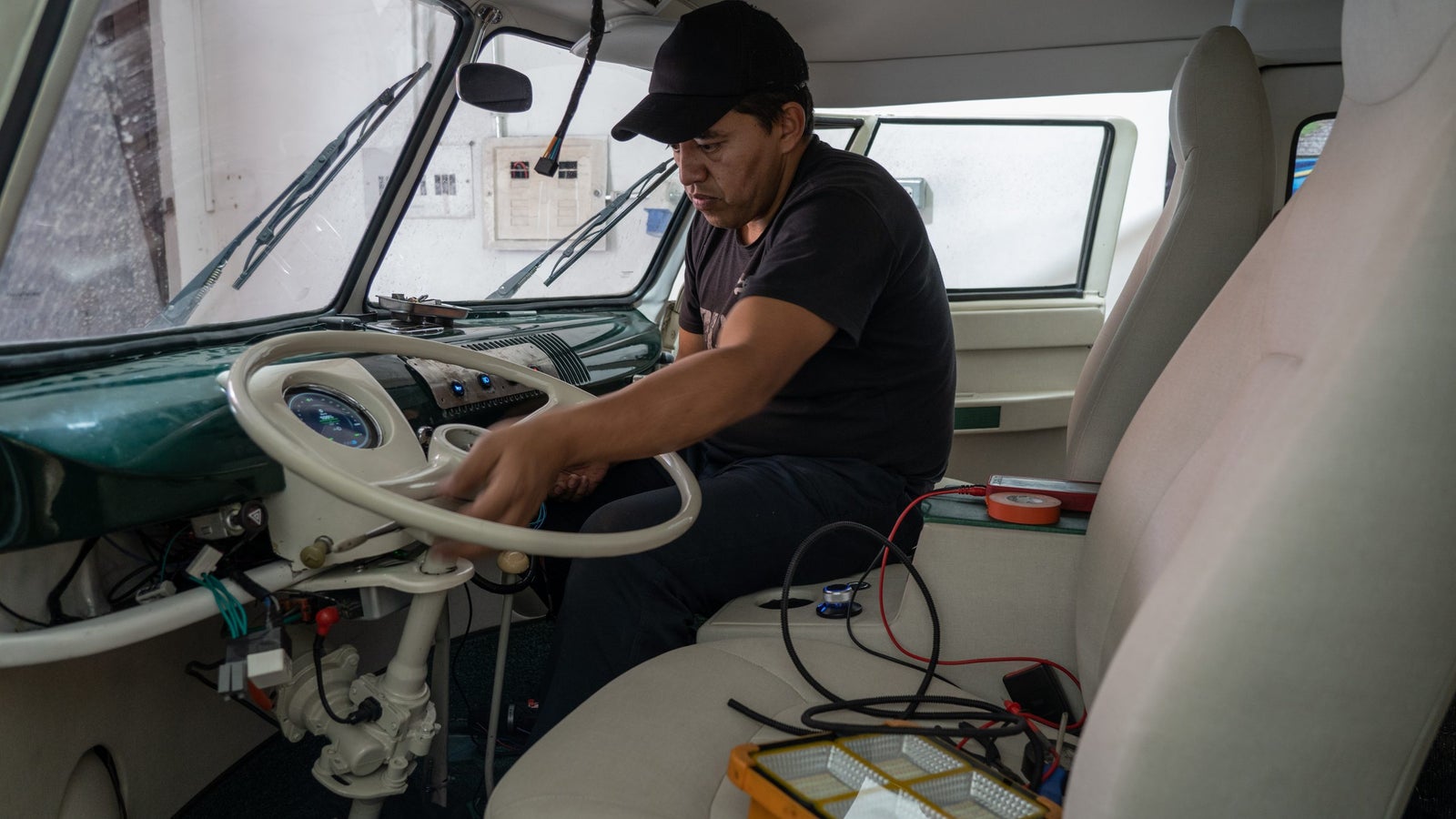 A photograph of a car mechanic working on a steering wheel inside a car. On the seat are many cables and reapair components.