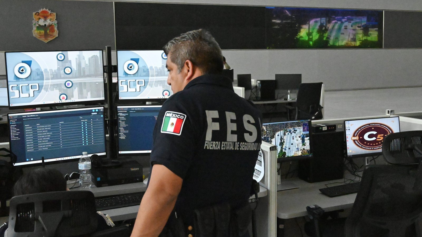 A photo of a Mexican official inside an office looking at a setup of four screens. There is a sign in the back that says C4 Baja California.