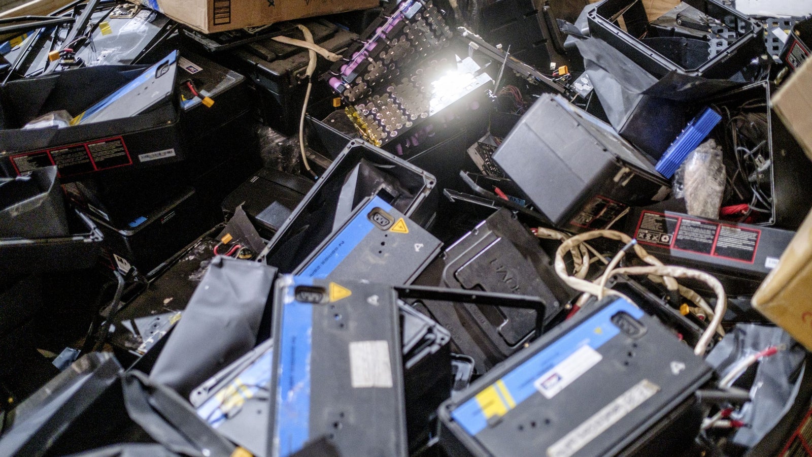 A chaotic pile of discarded batteries and battery casings scattered across a surface, with some cables and a cardboard box partially visible in the background.