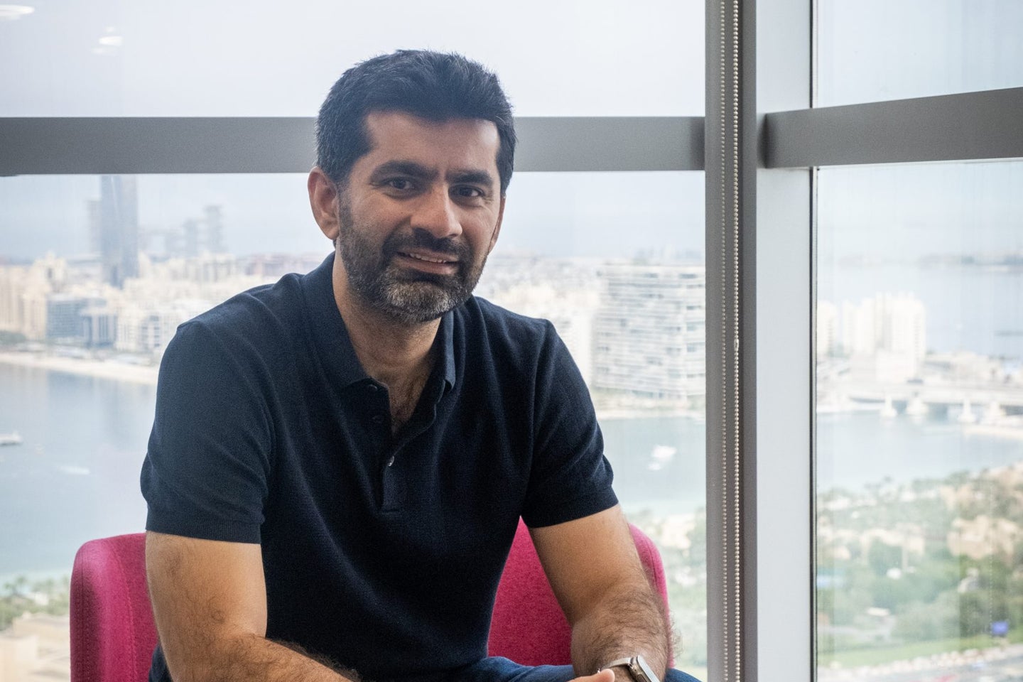 A man with a beard is seated on a pink chair next to a wooden side table, smiling and resting his hands on his knees. He is wearing a black polo shirt and blue jeans, with a city skyline and waterfront visible through the large window behind him.