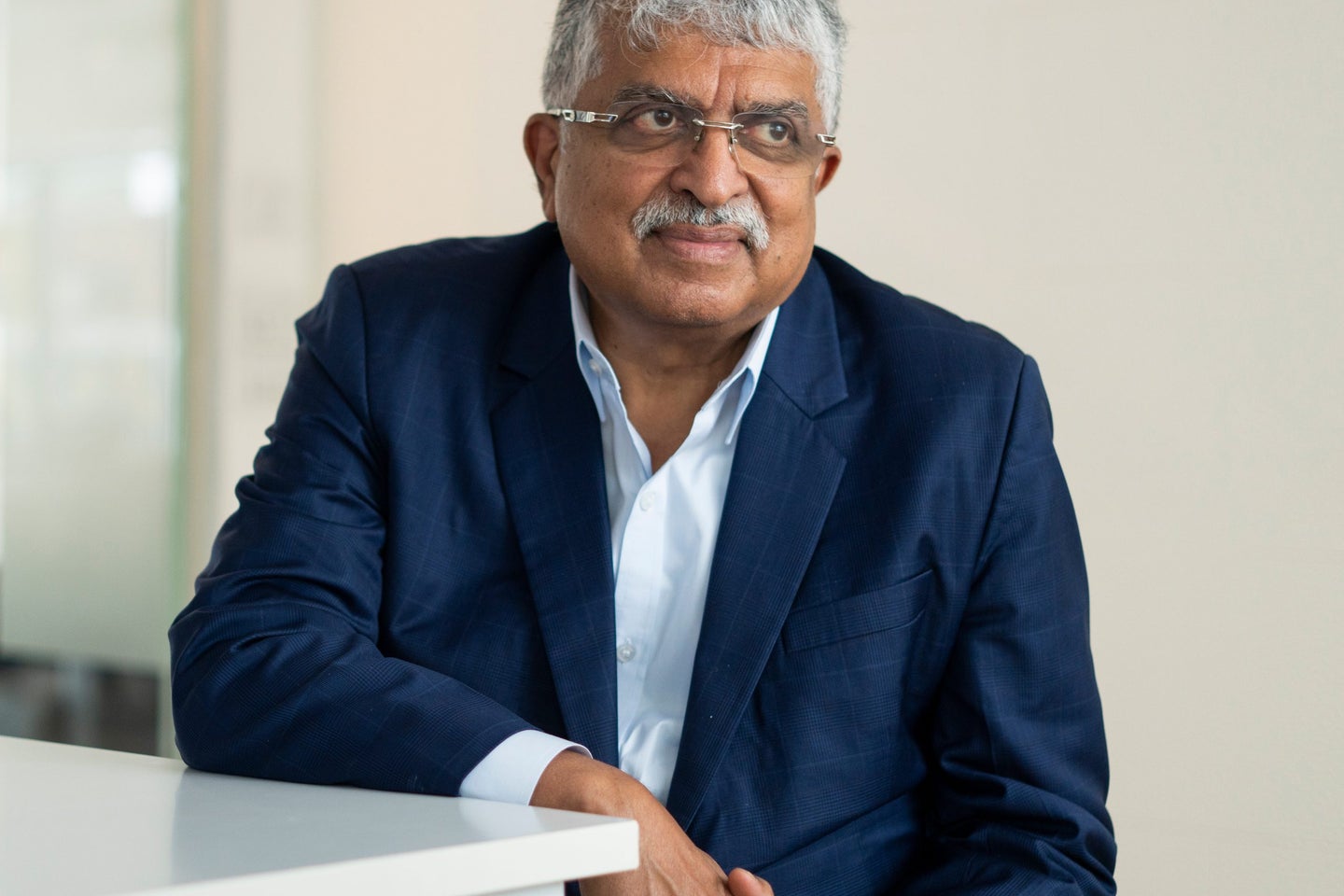 Nandan Nilekani, CEO of Infosys, with gray hair and glasses is seated at a table, wearing a blue blazer over a white shirt, looking thoughtfully to the side in a well-lit, modern environment.