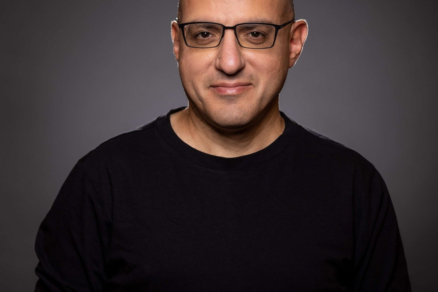 A bald man with glasses wearing a black long-sleeve shirt poses for a portrait against a dark background, smiling slightly.