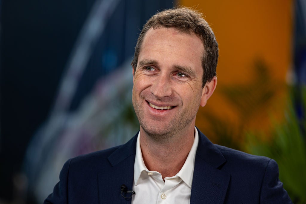 Man with short brown hair and a slight beard, smiling while wearing a dark suit and white shirt, seated in front of a blurred background with hints of greenery and colorful graphic elements.