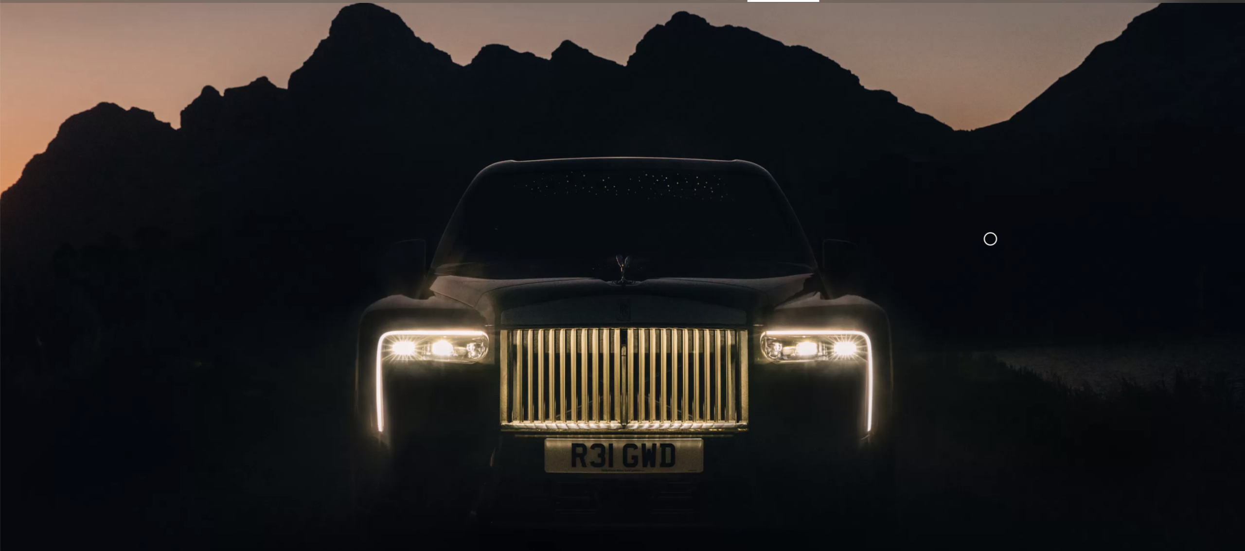 Главный, темный роскошный автомобиль с выдающейся передней решеткой и сияющими фарами расположен на фоне буровых гор в сумерках, подчеркивая его элегантный дизайн при слабом освещении.