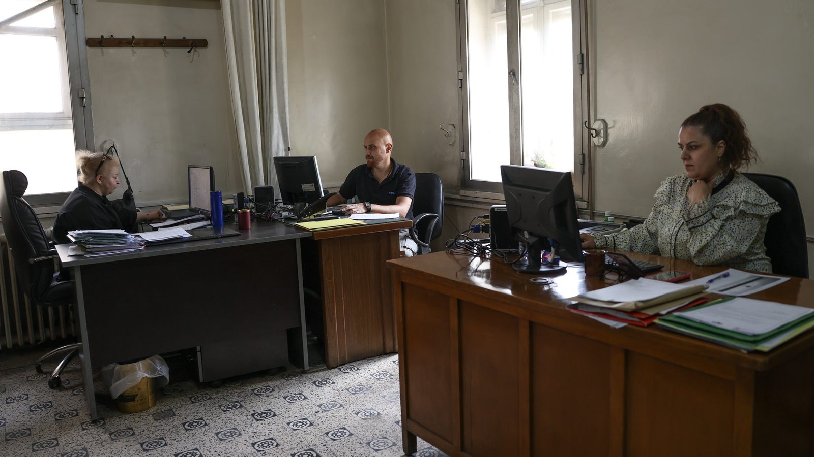 Three people working in an office; one woman at a desk on the left using a computer, a man in the center working on his desk, and another woman on the right focused on her computer, surrounded by paperwork and file folders.