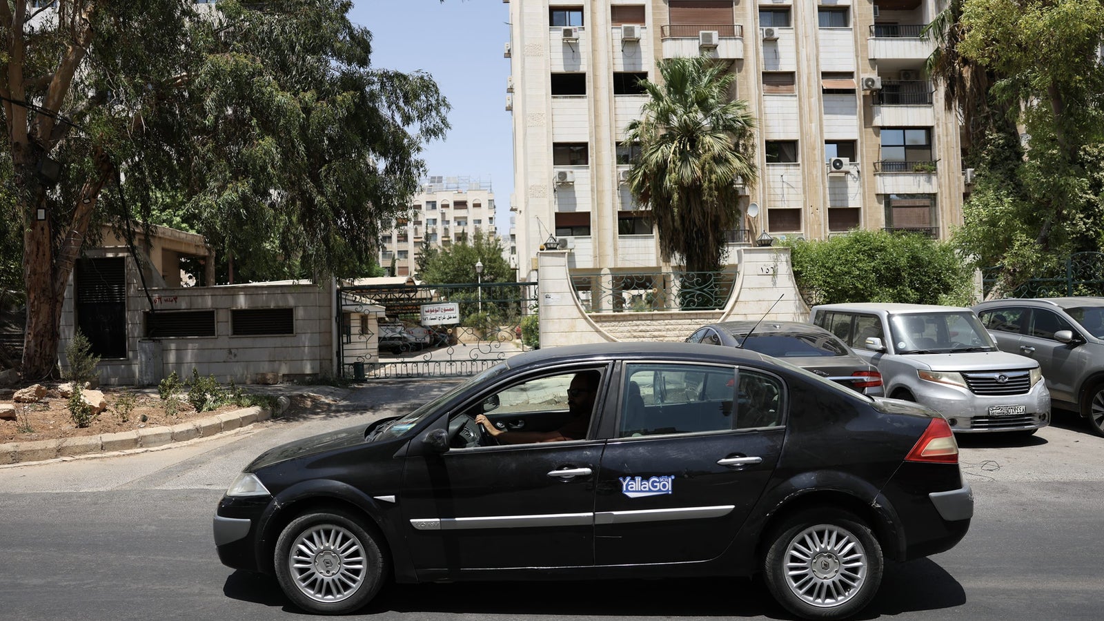 A black car with a 'Yalla Go' sticker drives on a street lined with trees and residential buildings. In the background, there are multiple parked cars and a gated entrance to a building complex.
