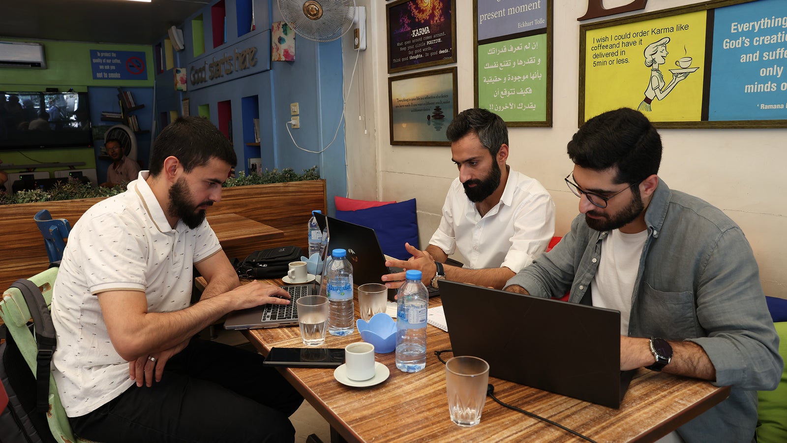Three men are working at a cafe table, each using a laptop. One man in a white shirt is focused on his screen while gesturing with his hands. Another man in a grey shirt appears to be typing, and the third man, wearing glasses, is looking down at his laptop. The table is cluttered with cups, glasses of water, and a small decorative item, with colorful cushions and wall art visible in the background.