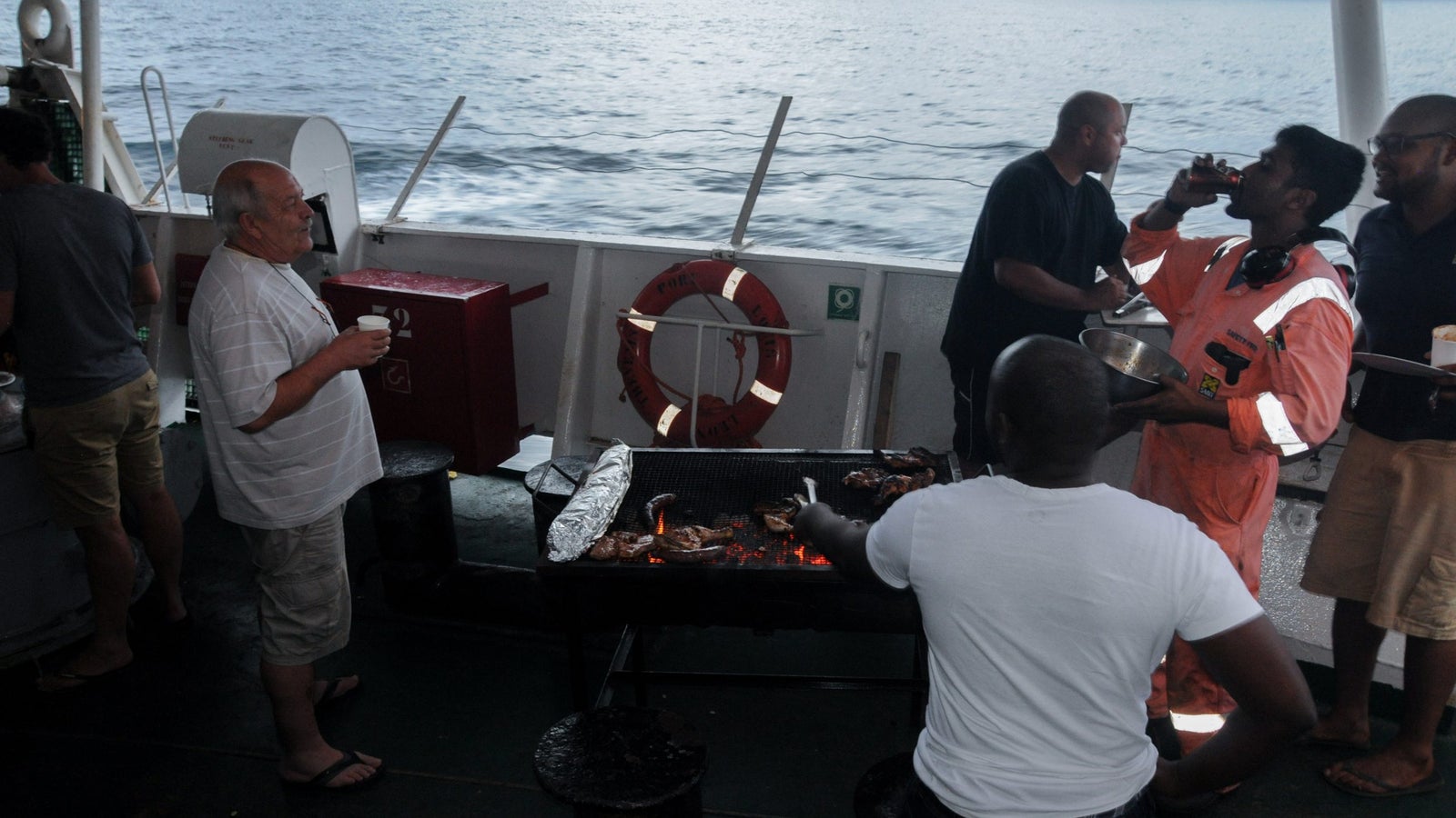 A group of men barbecuing on a boat, with one man in an orange jumpsuit holding a drink and another grilling chicken. The background features calm ocean water and a lifebuoy.