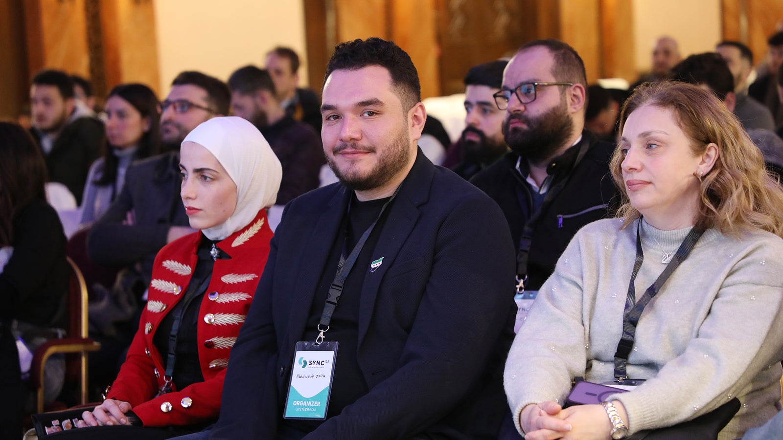 A diverse group of attendees sitting in a conference setting, with a woman in a red military-style jacket and white hijab, a smiling man in a dark blazer with a badge, a woman in a gray sweater with a patterned design, and others in the background, all focused on the event.
