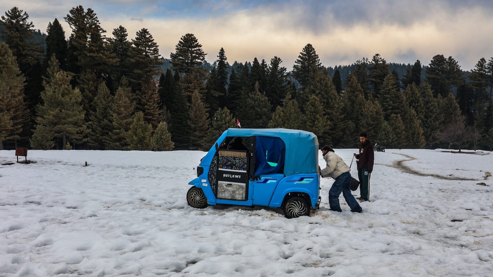 Two men help push a blue auto rickshaw stuck in snow, surrounded by a snowy landscape and pine trees in the background.