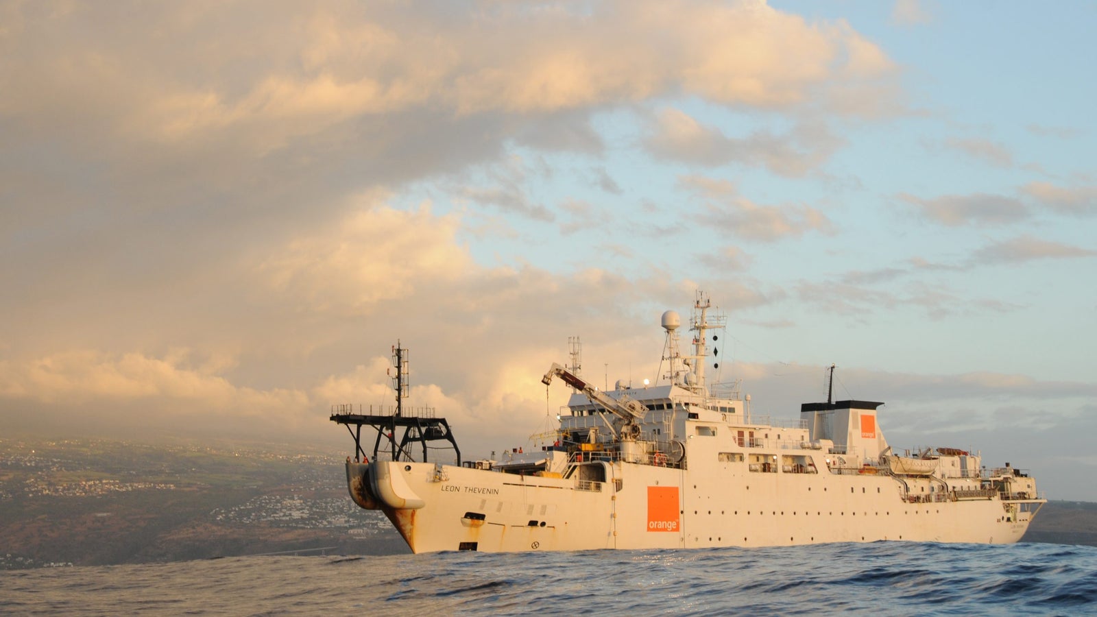 A research vessel named 'León Thévenin' is situated on the ocean with a backdrop of clouds and a distant coastline, showcasing its scientific equipment and features against a vibrant sky.