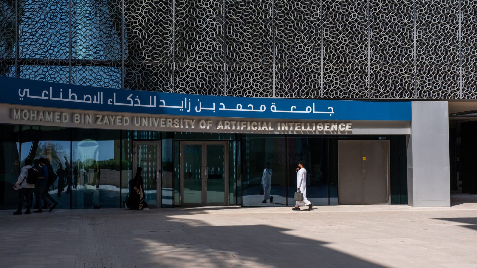 The entrance of the Mohamed Bin Zayed University of Artificial Intelligence, featuring modern architecture with intricate geometric patterns, blue signage, and a reflection on the glass doors. A person dressed in traditional attire walks by.