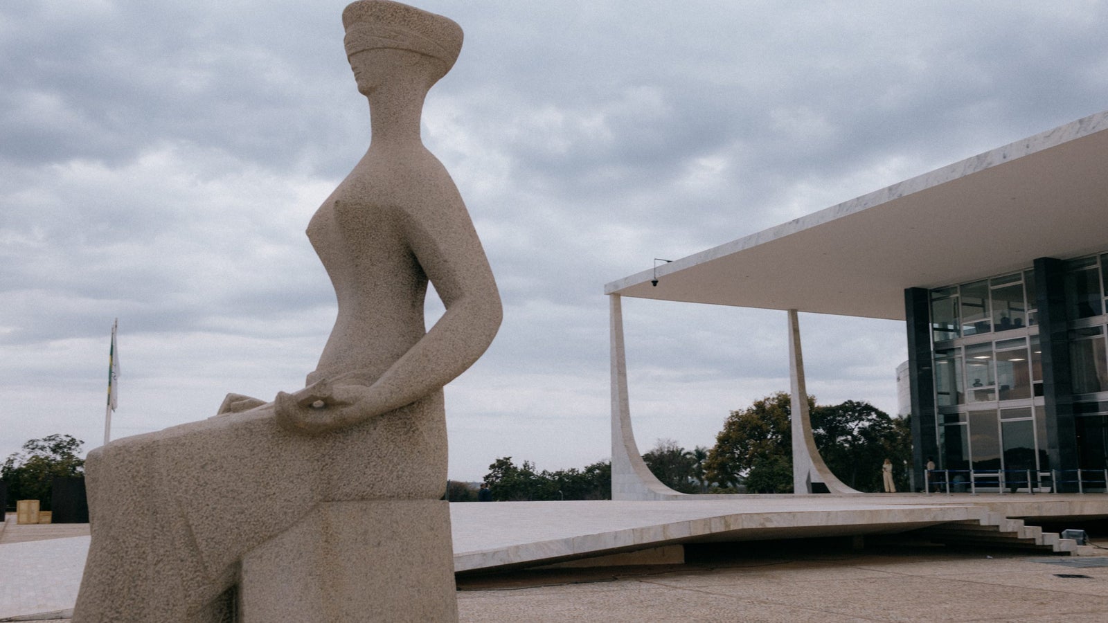 A large stone sculpture of a blindfolded woman seated with a serene expression, situated in an open plaza in front of a modern architectural building with a flat roof and large glass windows, under a cloudy sky.