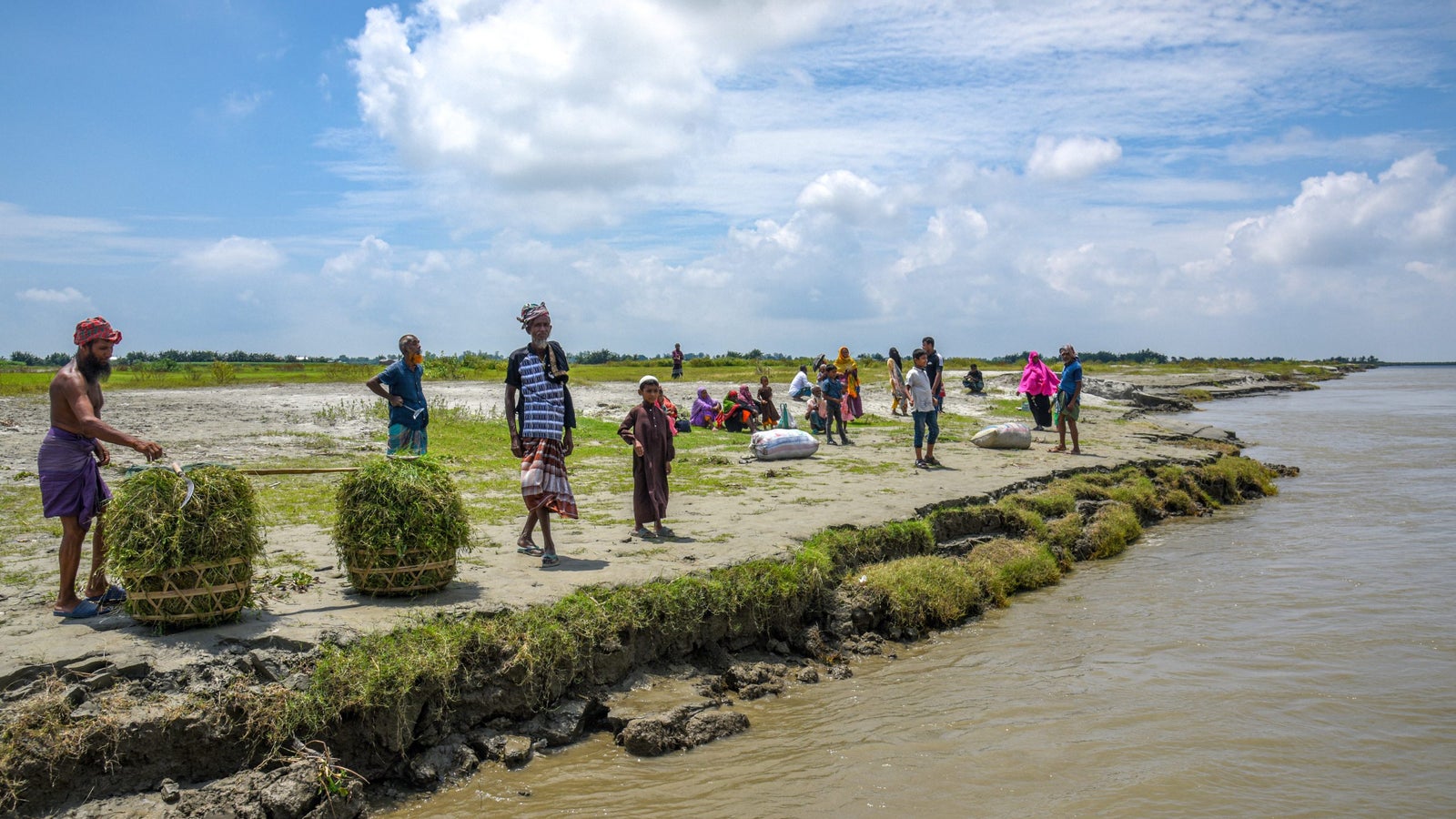 A group of people gathers on a riverbank under a partly cloudy sky, with one man transporting bundles of grass on a cart, while others are engaged in various activities. The scene includes men, women, and children, with some wearing traditional clothing and sitting on the sandy ground, surrounded by greenery and the flowing river.