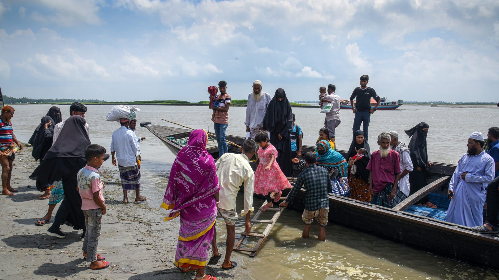 A diverse group of people gathered at a riverside preparing to board a boat, with children, men, and women in traditional clothing. Some carry bags and children, while others assist with loading. The background features a cloudy sky and greenery across the water.