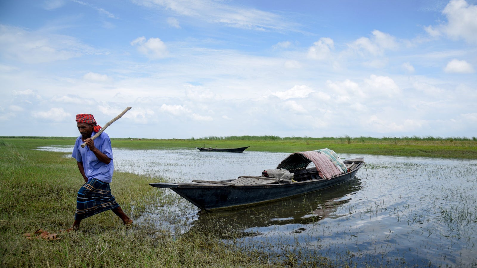 A man in a blue shirt and traditional patterned wrap walks through shallow water carrying a stick, with grassy land and two wooden boats in the background under a partly cloudy sky.