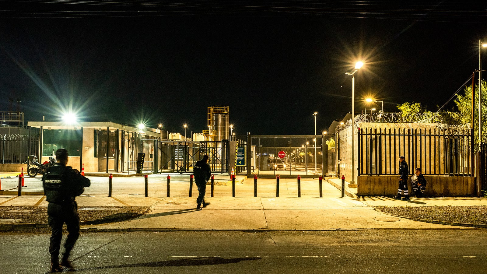 Night scene of a secure facility with armed guards patrolling near a barbed-wire fence and bright floodlights.