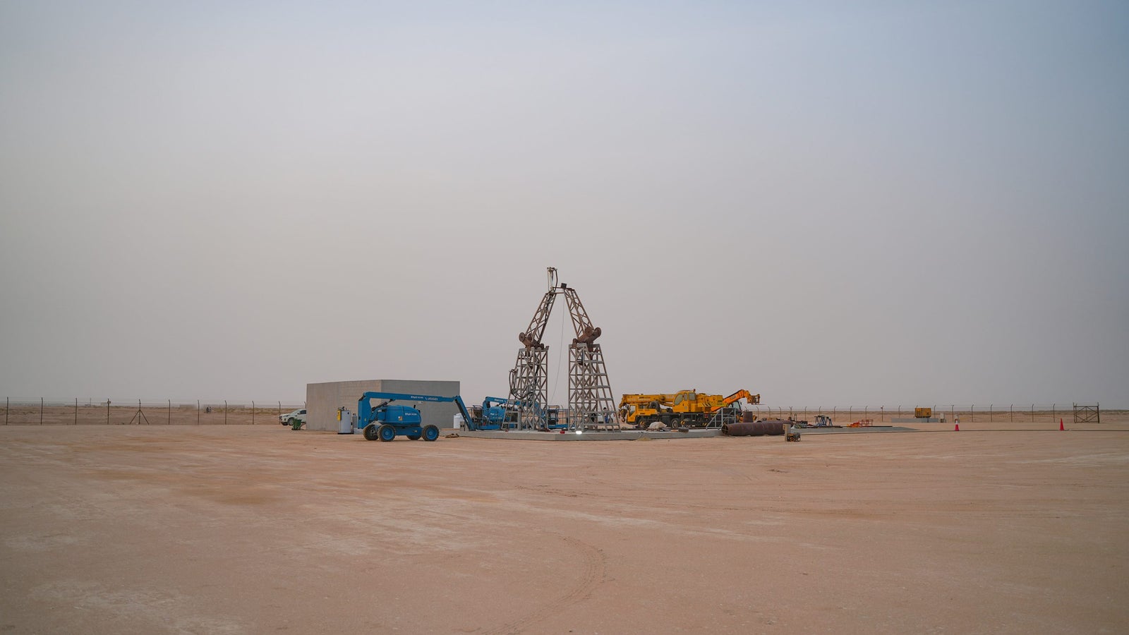 A large, industrial construction site in an arid landscape, featuring a tall, metallic structure resembling a crane with two triangular frames. Various construction vehicles, including blue lift machines and a yellow truck, are scattered around a gray concrete block structure on the sandy ground, with a hazy sky and fencing in the background.