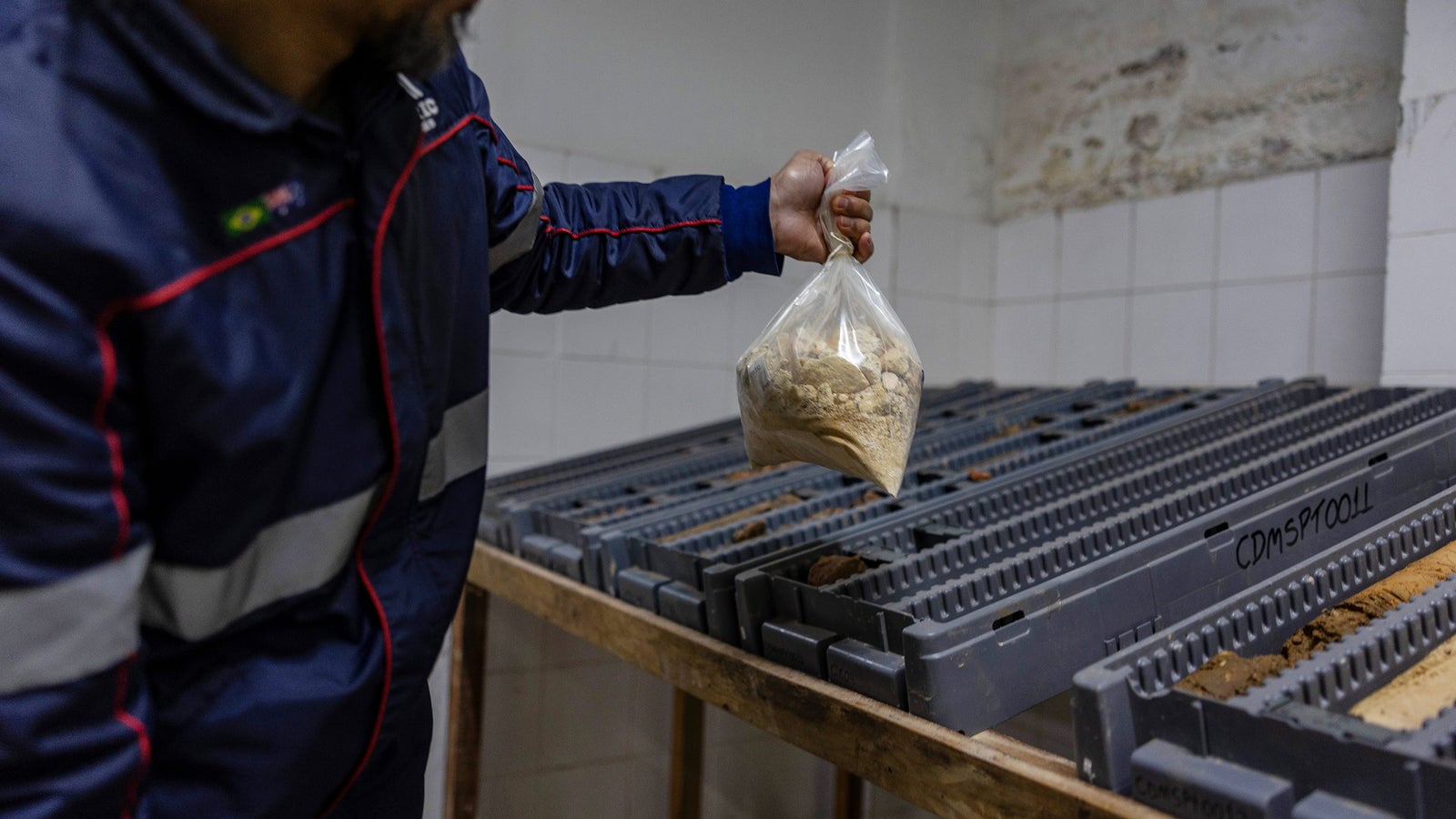 A person wearing a dark blue jacket with reflective stripes holds a plastic bag containing a light brown substance, while standing beside a wooden table with gray trays arranged in rows, in a room with white tiled walls.
