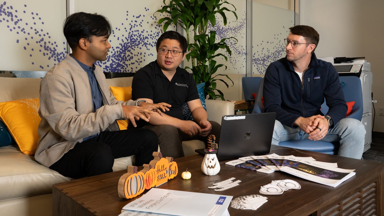 Three men are engaged in discussion on a couch with colorful pillows, a laptop, and festive decorations on a coffee table.