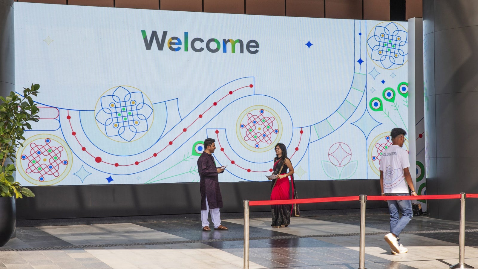 A welcome display with two people chatting in traditional attire and a man in a volunteer shirt walking past.