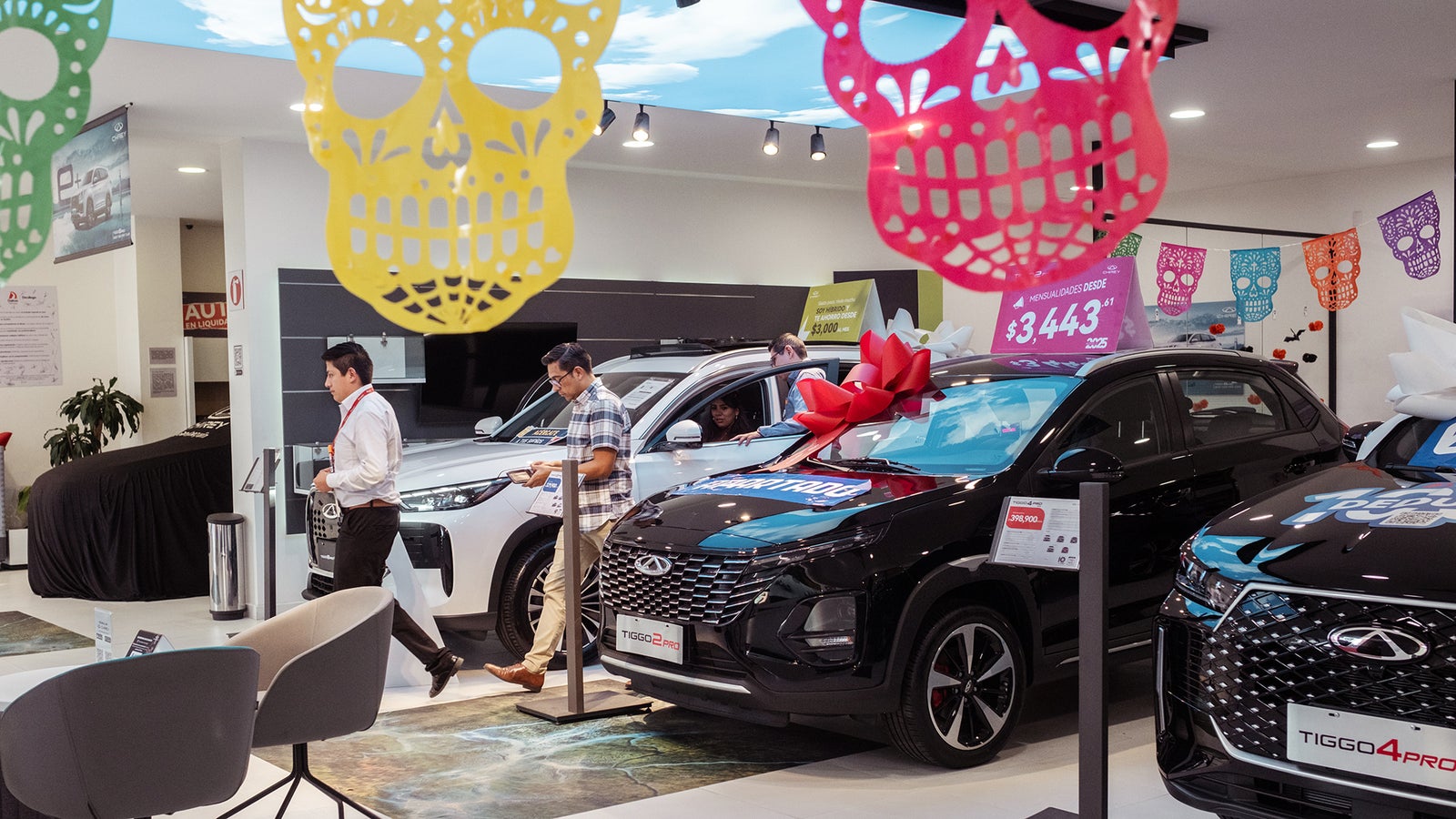Inside a car dealership, men browse vehicles decorated with bows and colorful paper decorations for a festive theme.
