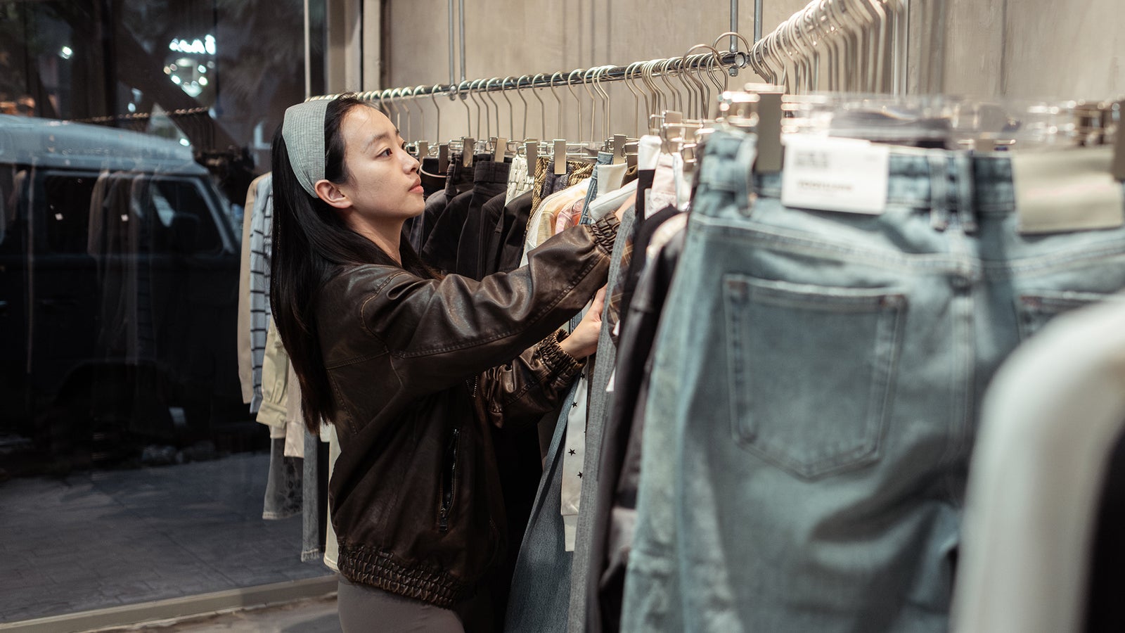 A woman with long hair in a dark jacket browses clothing on hangers in a store.
