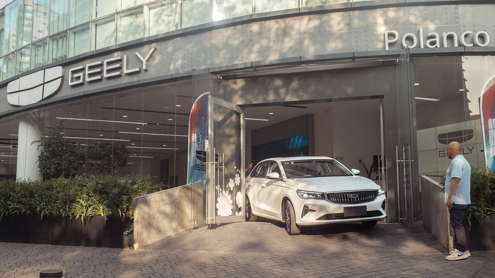 A white Geely car is driving out of a modern dealership, with the Geely logo visible and a man observing nearby.