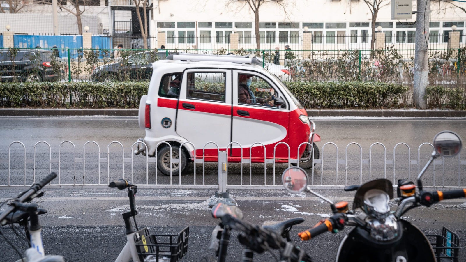 A small, red and white electric vehicle drives past a row of parked bikes along a city street with trees in the background.