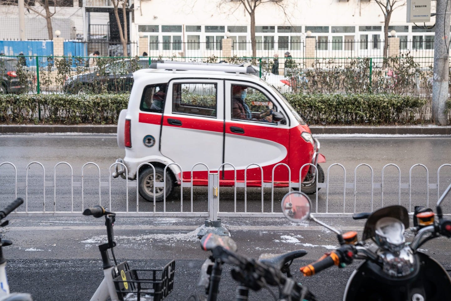 A small, red and white electric vehicle drives past a row of parked bikes along a city street with trees in the background.