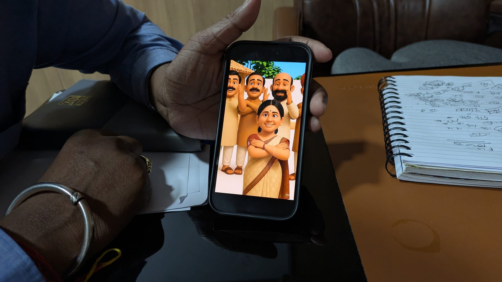 A hand holding a smartphone displaying animated characters: a woman in a sari and several smiling men behind her. A notepad is nearby.