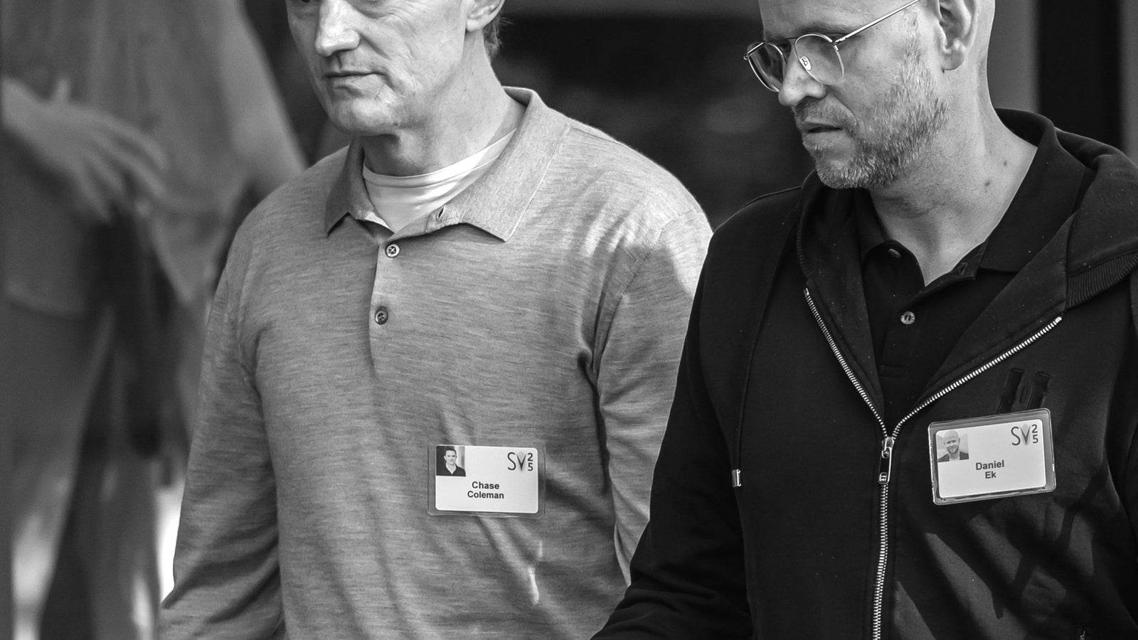 Two men walking together; one wears a gray polo shirt and the other a black hoodie, both with name tags.