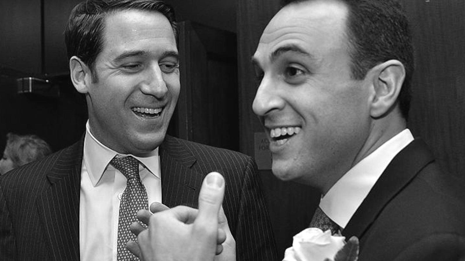 Two men in suits smiling and interacting in a dimly lit room, one wearing a flower in his lapel.