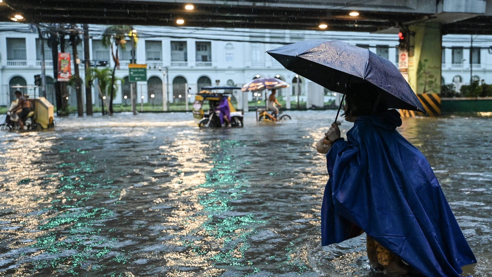A person in a blue raincoat holding an umbrella walks through a flooded street with standing water and vehicles in the background.