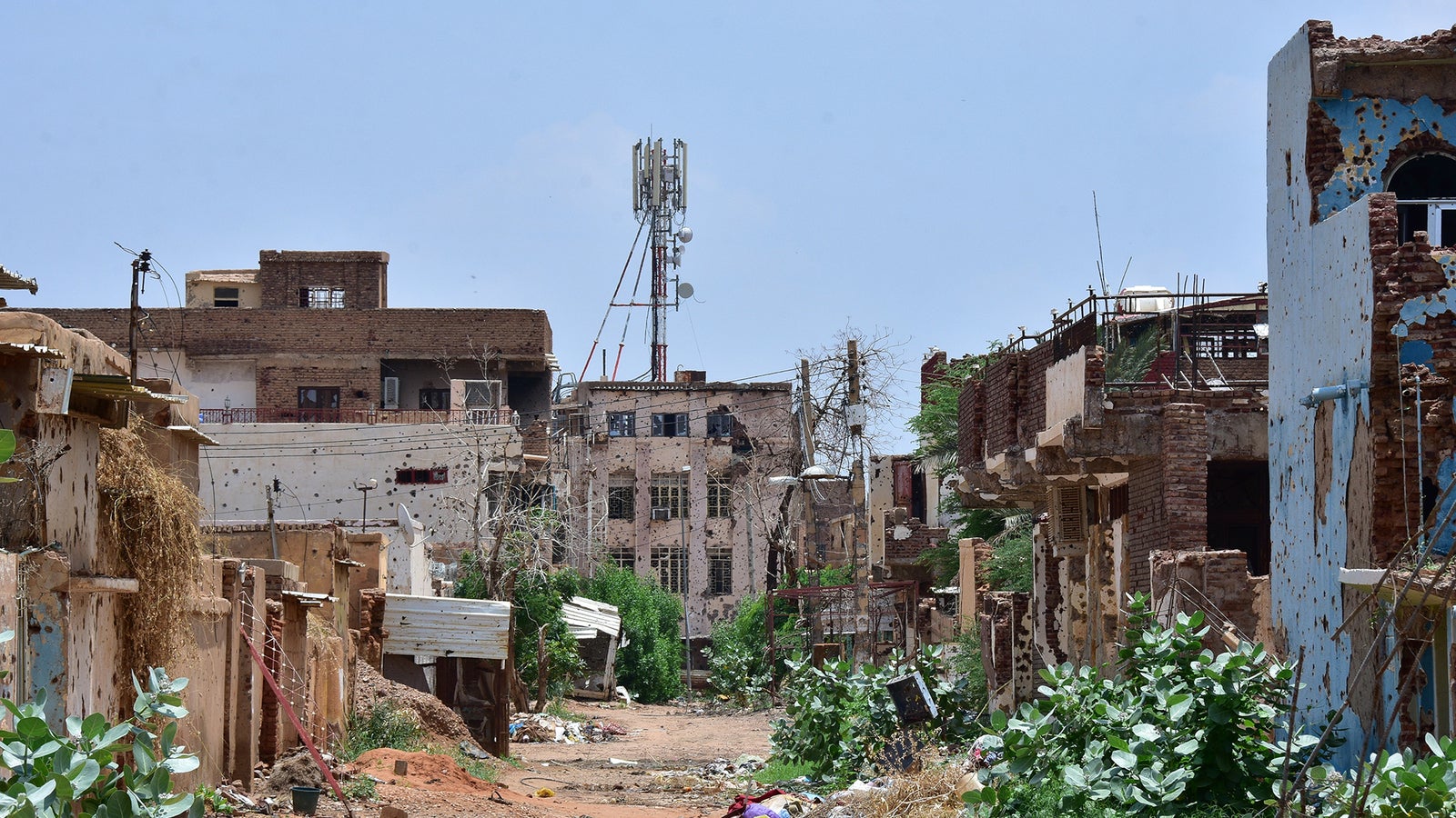 A deserted street in a dilapidated area, featuring crumbling buildings, overgrown vegetation, and a cell tower in the background.