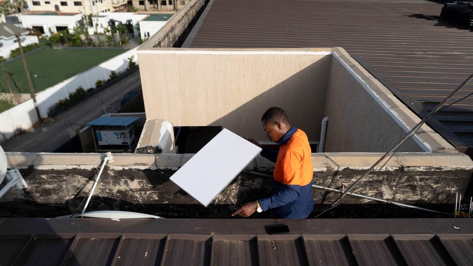 A worker in an orange and navy uniform carries a large panel across a rooftop, with buildings and a sports field in the background.
