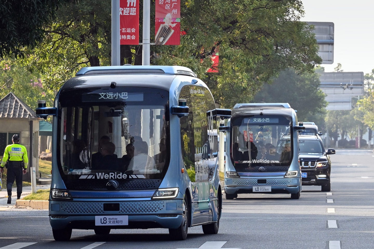 Two autonomous buses are driving down a city street, with trees and buildings visible in the background.