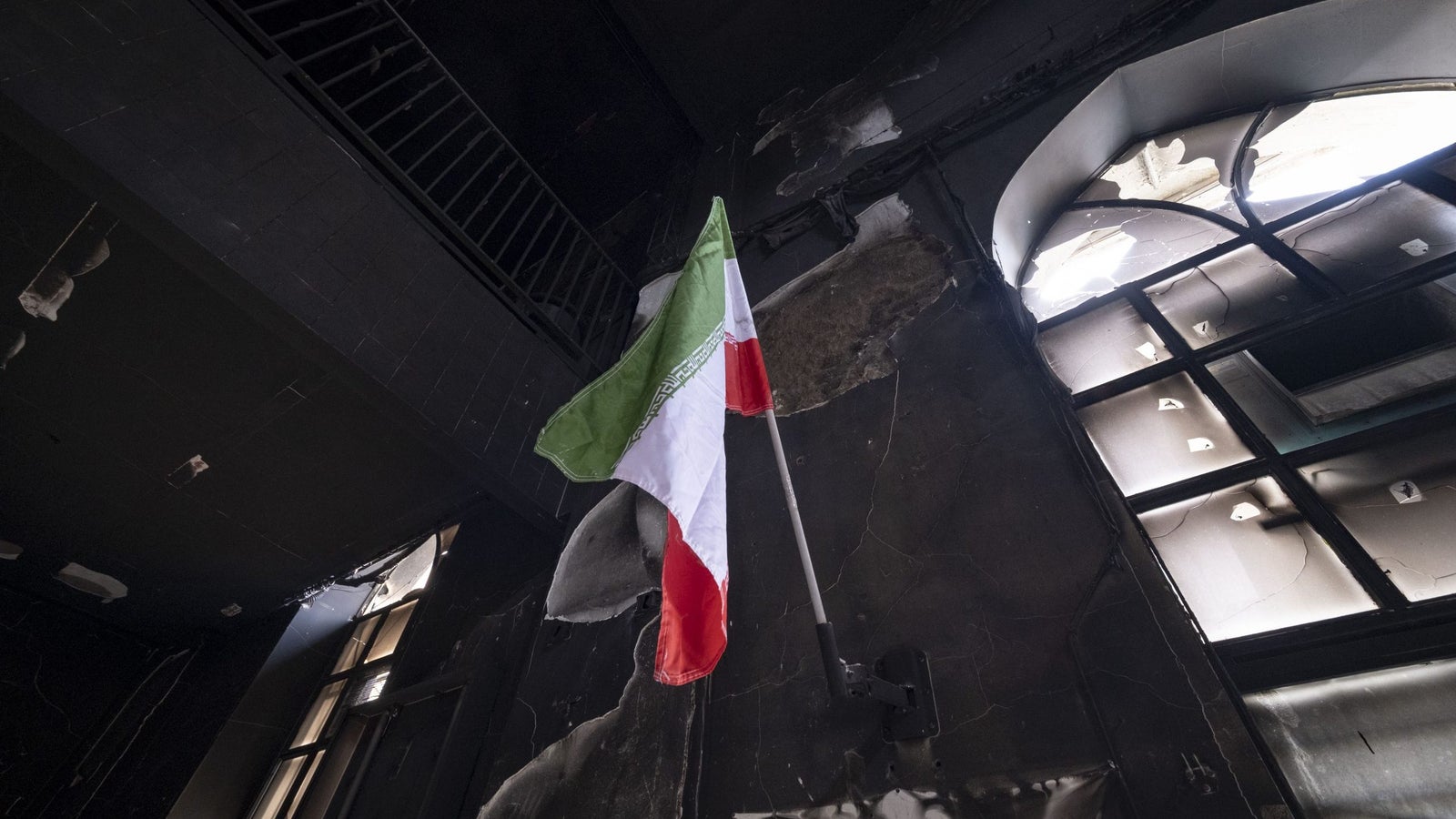 A tattered Italian flag hangs in a charred, darkened room with damaged windows and walls.