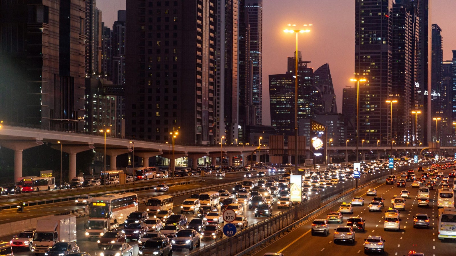 Busy highway scene at dusk, filled with vehicles and illuminated by streetlights, with tall city buildings in the background.