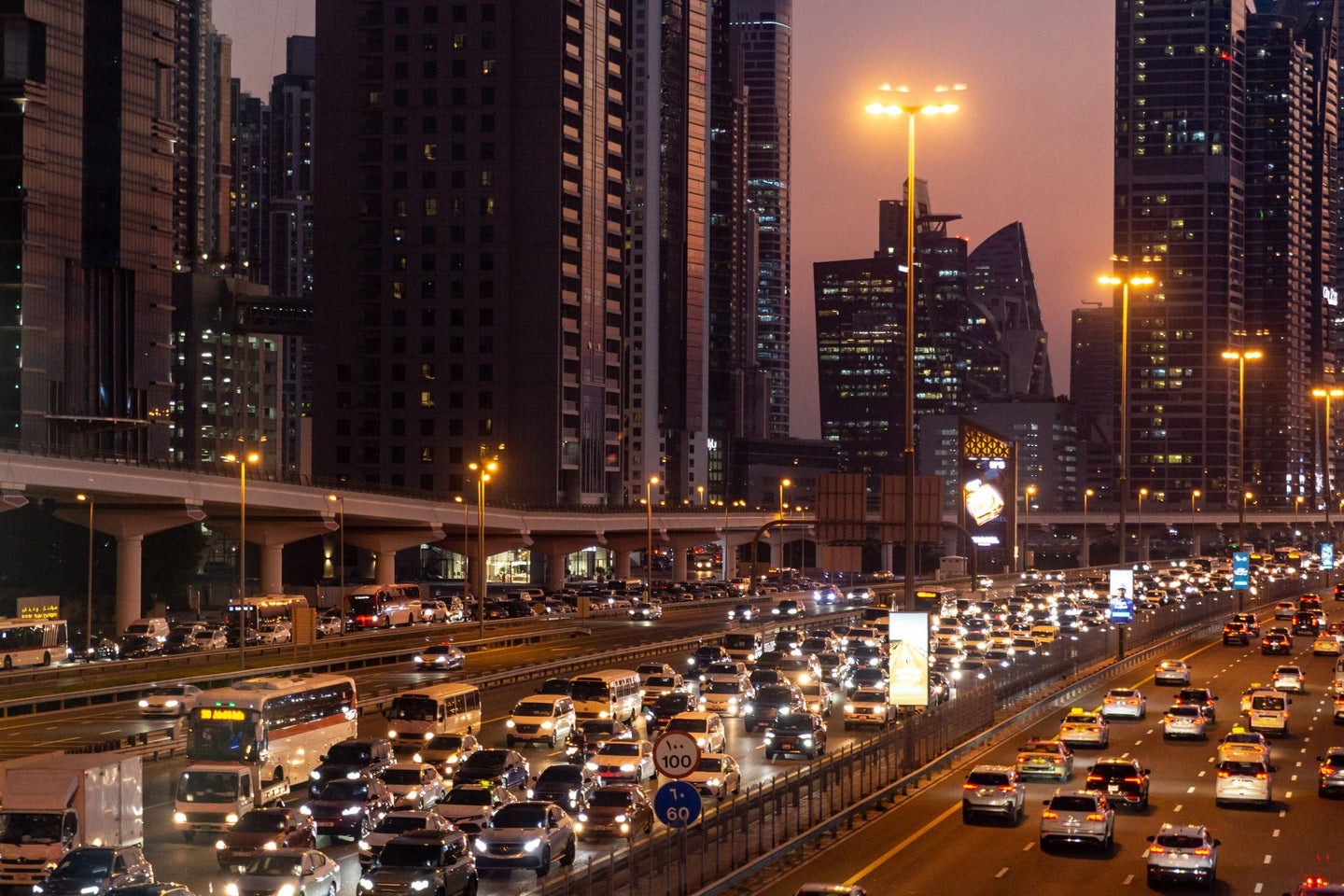Busy highway scene at dusk, filled with vehicles and illuminated by streetlights, with tall city buildings in the background.