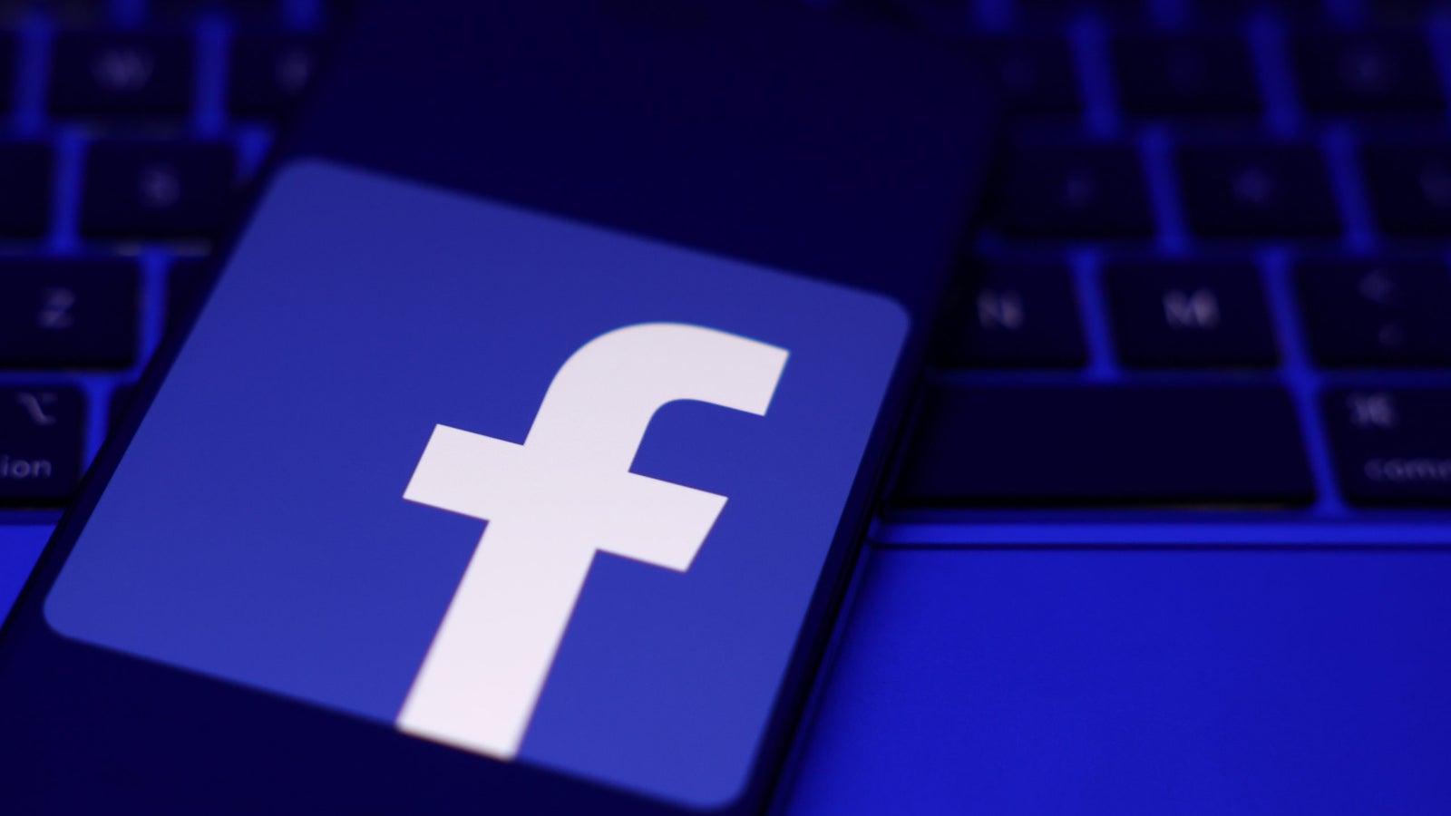 A smartphone displaying the Facebook logo, placed on a dark keyboard with a blue hue.