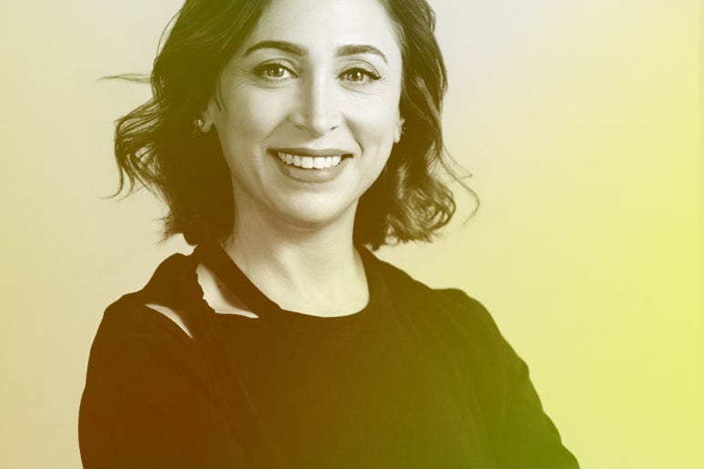 A woman with shoulder-length wavy hair smiles confidently, wearing a black shirt against a softly colored background.